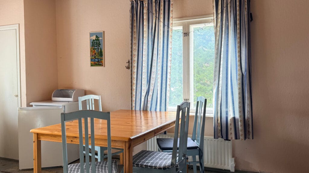 Ikkunan edessä pöytä, jonka ympärillä neljä siniharmaata tuolia. Ikkunassa sinivalkoiset verhot.