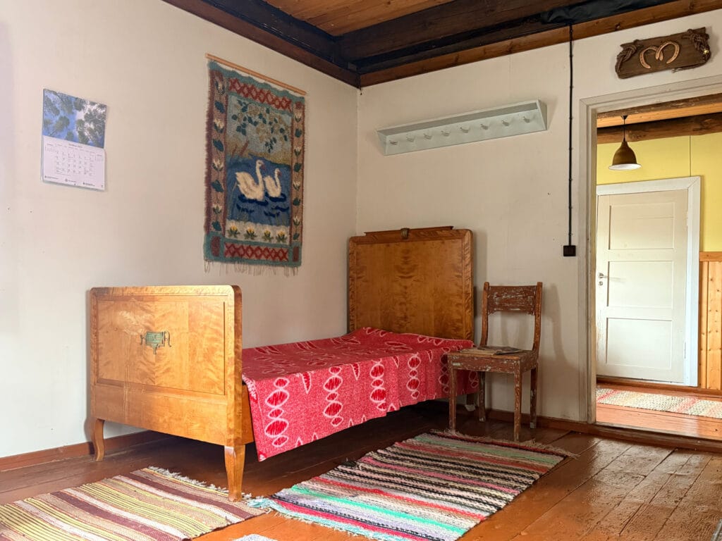 Huone, jossa lankkulattia. Huoneesta näkyy nurkka, johon on sijoitettu puinen sänky. Sängyn yläpuolella ryijy, jossa kuva joutsenista. Sängyn oikealla puolella oviaukko