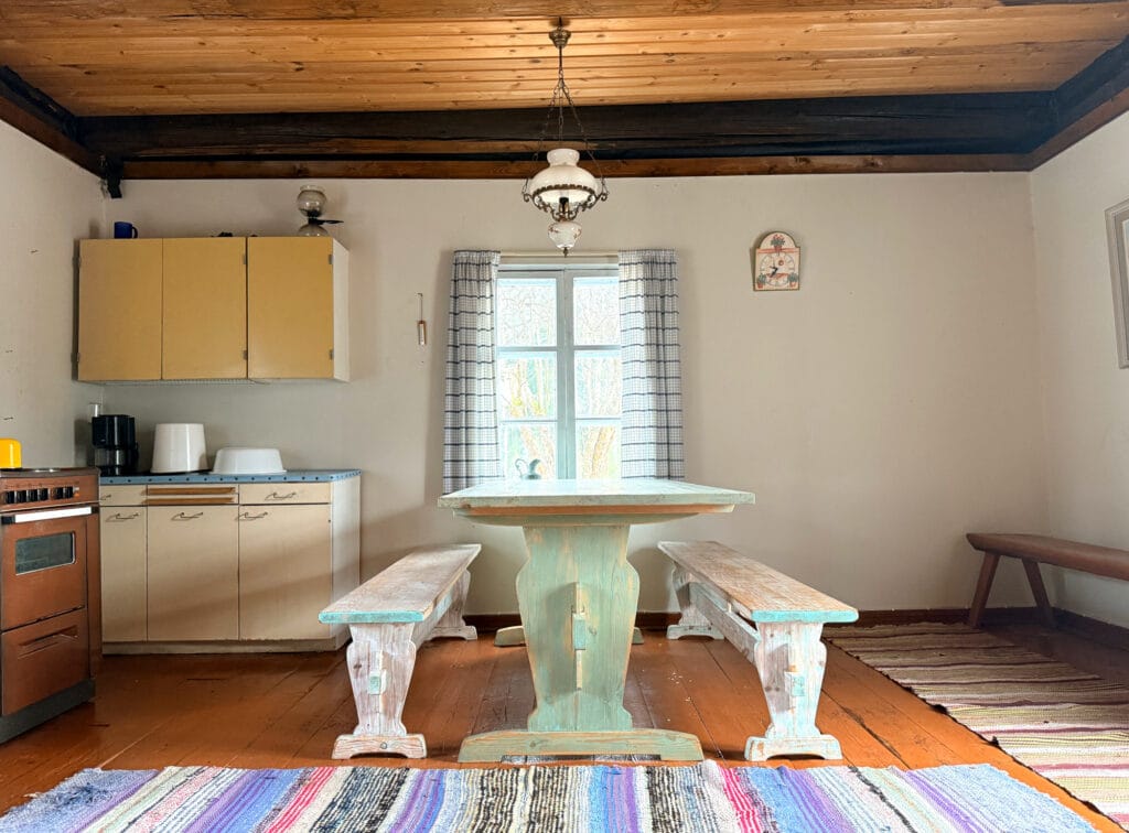 Vasemmalla liesi ja tiskikaappi. Keskellä kuvaa pirtinpöytä, jonka takana ikkuna, jossa ikkunaruudukko ja sinivalkoruutuiset verhot. Lattialla räsymatot.