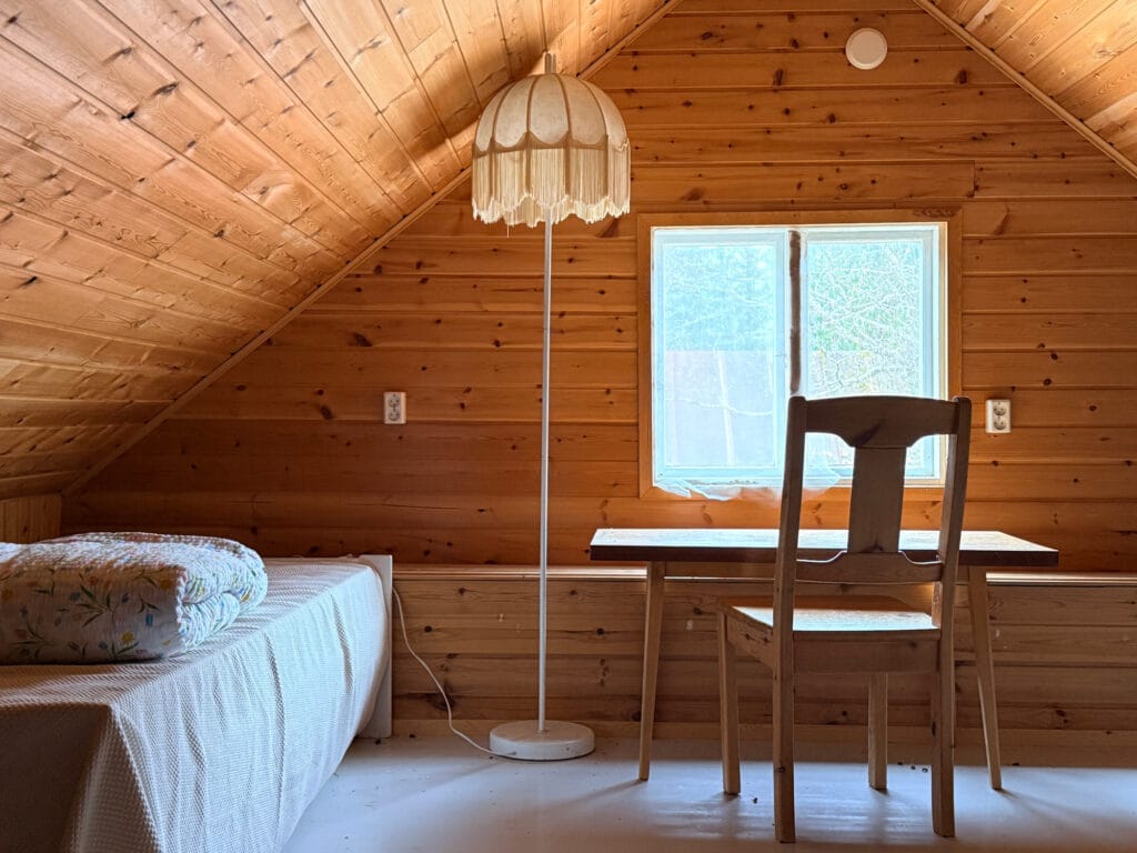 Huone, jossa vasemmalla puolella yhden hengen sänky, vieressä korkea jalkalamppu. Ikkuna, jonka edessä kirjoituspöytä ja tuoli.