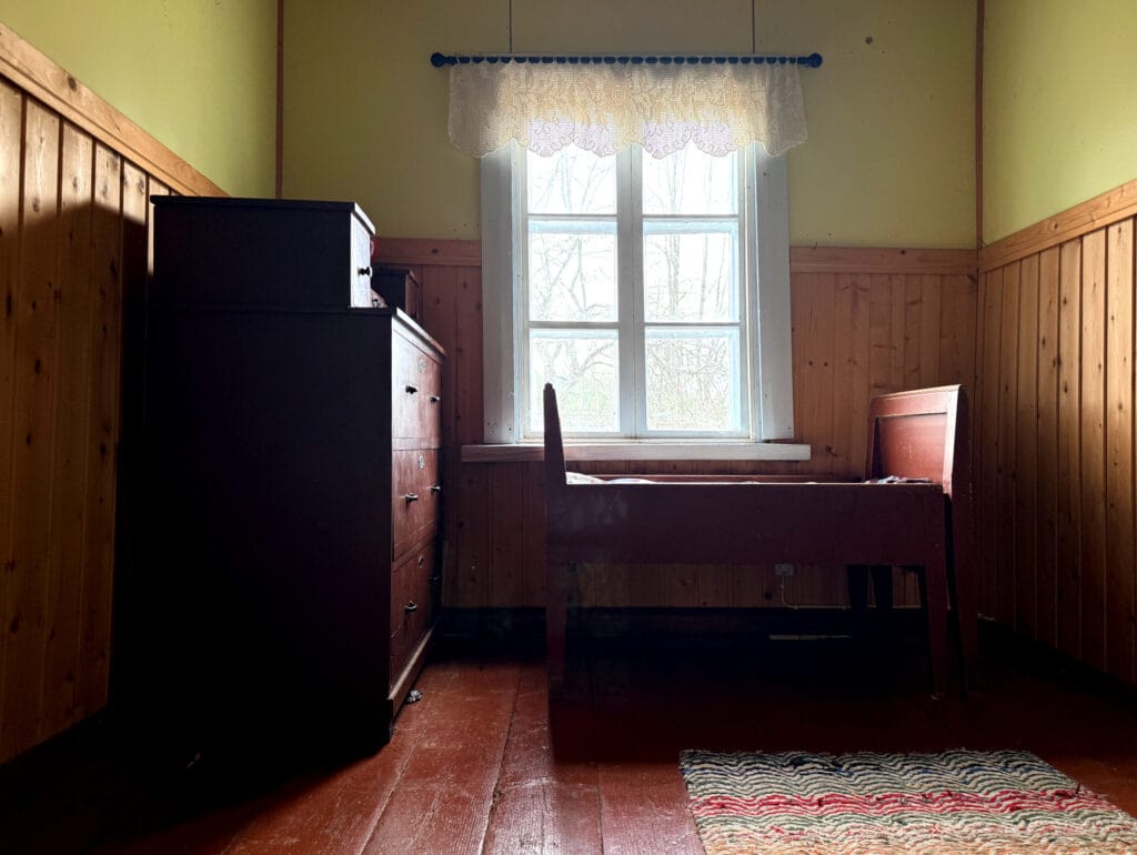 Huone, jossa ikkunan edessä puinen sänky ja lipasto. Lattia lankkua ja räsymatto
