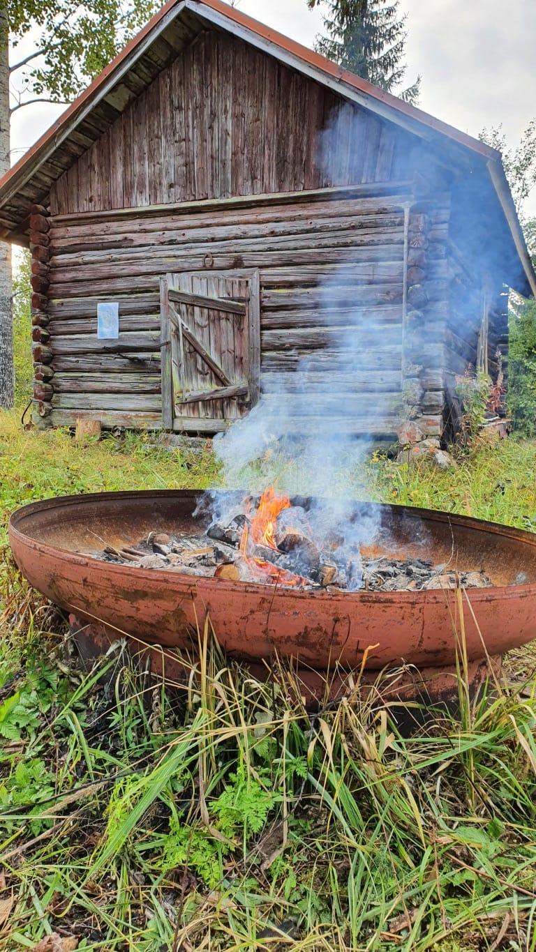 Savuava ulkotuli, jonka takana vanha harmaantunut hirsirakennus.