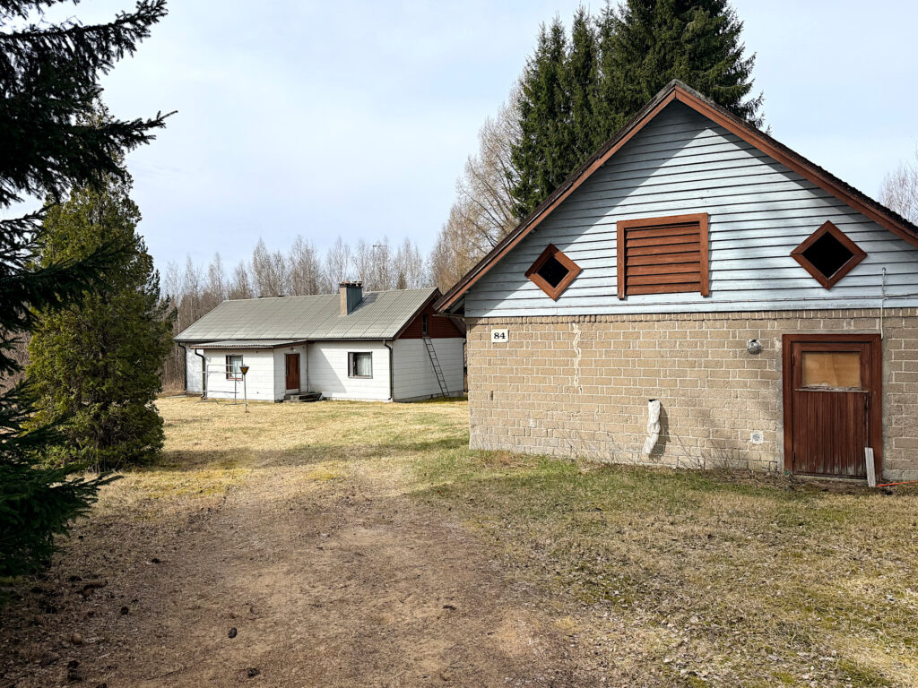 Oikeassa reunassa kaksivärinen talonpääty, jossa ruskeat ikkunat ja ovi. Takana harjakattoinen vaalea päärakennus