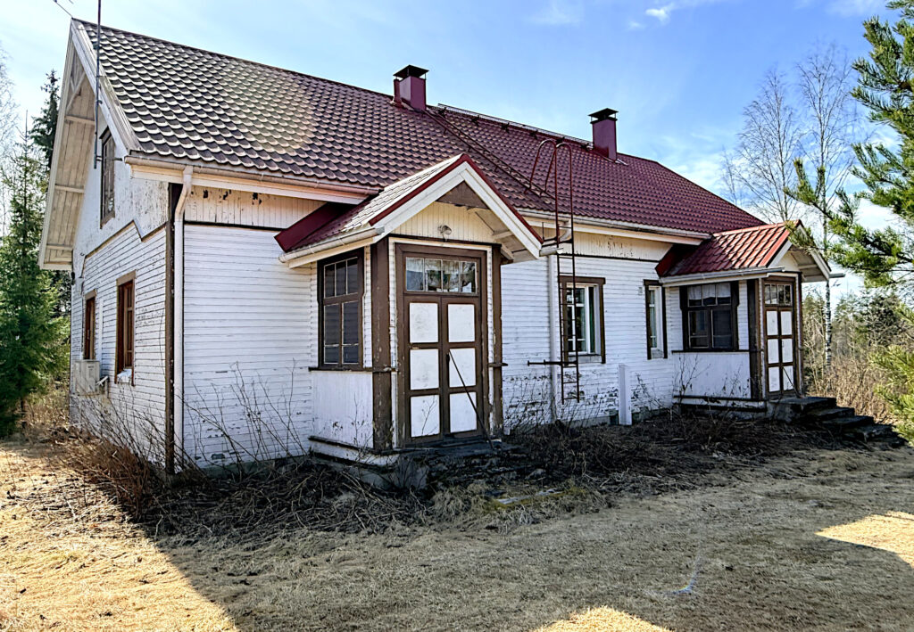 Vaalea iso puurakennus, punainen katto ja kaksi kuistia.