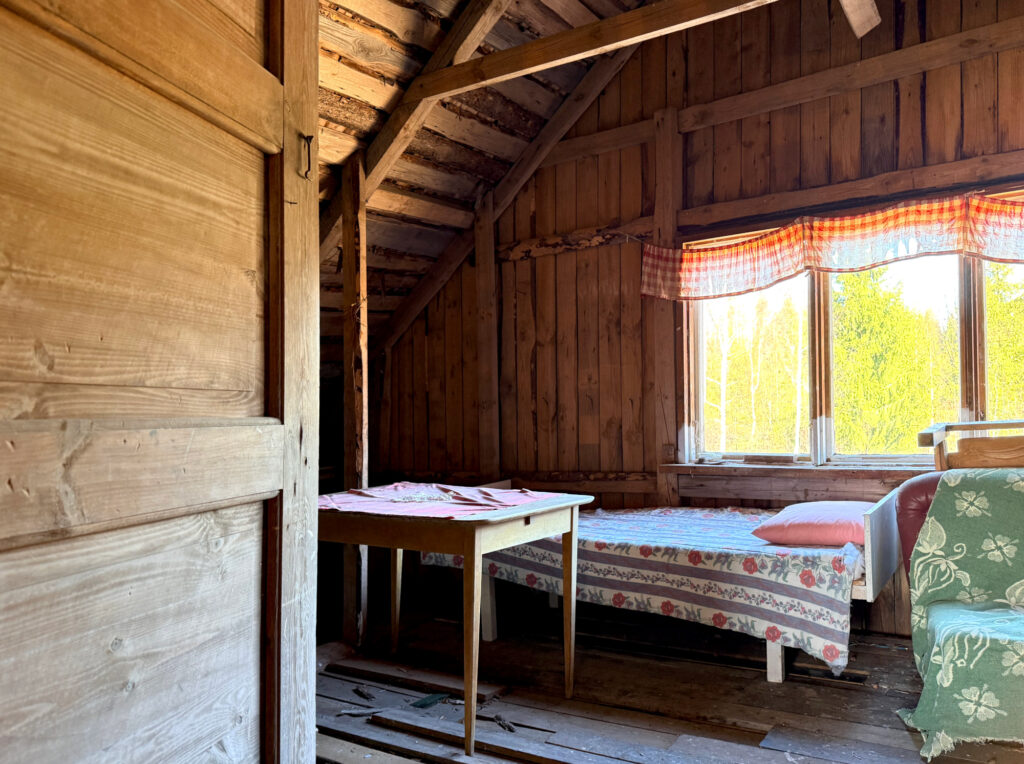 Näkymä ovelta kesähuoneeseen, jonka seinät ovat lautaa ja ikkunasta näkyy kesäinen maisema.