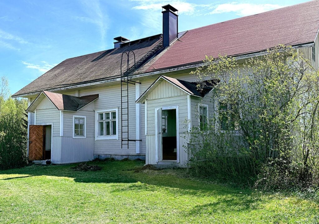 vaaleansävyinen talo kuvattuna ulkopäin. Punertava katto, vihreä ruoho, sininen taivas.