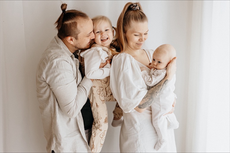 Perhe, jossa vanhemmat ja kaksi lasta. Kuvattuna hieman sivusta siten, että lapset ovat vanhempien sylissä. Sävy vaalea kuvassa ja perheen vaatteissa.