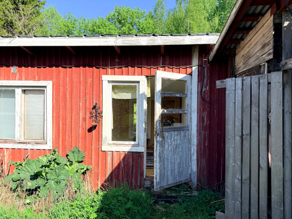 Punainen rakennus, jossa valkoinen ovi, jonka maali lohkeillut. Ovi raollaan.