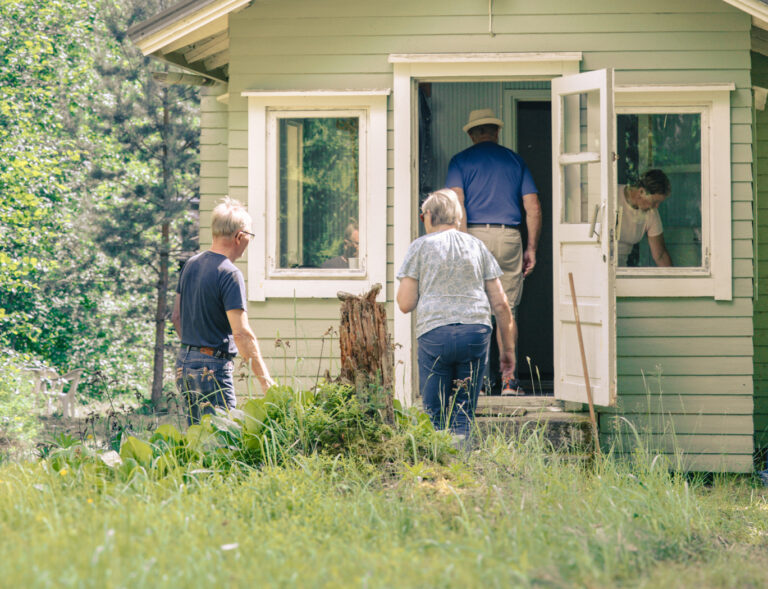 Vihreän puutalon kuistin edessä ja portailla ihmisiä siirtymässä sisälle taloon.
